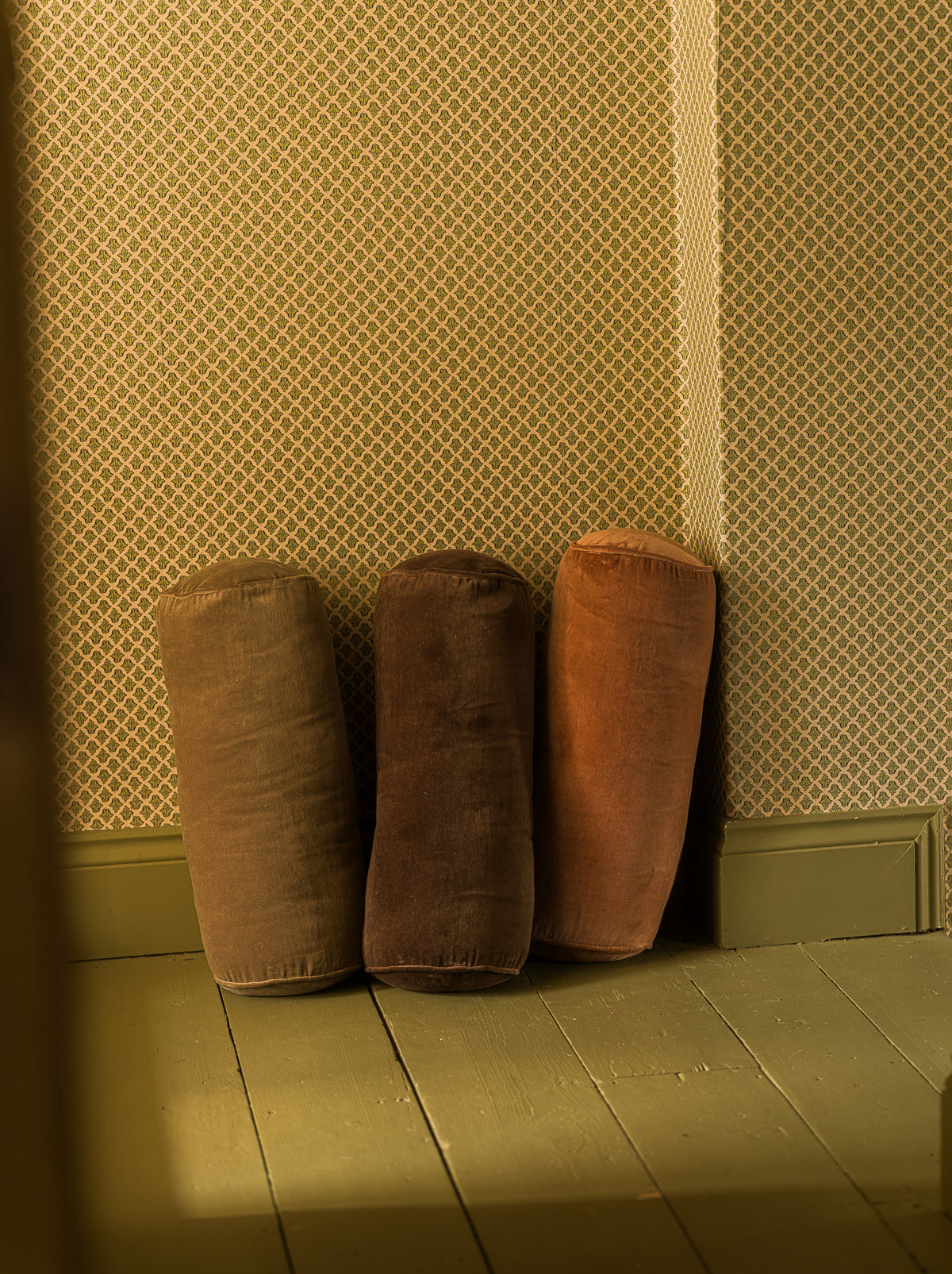 Three cylindrical floor cushions in brown and rust-orange velvet, arranged against a patterned wallpaper wall on a wooden floor.
