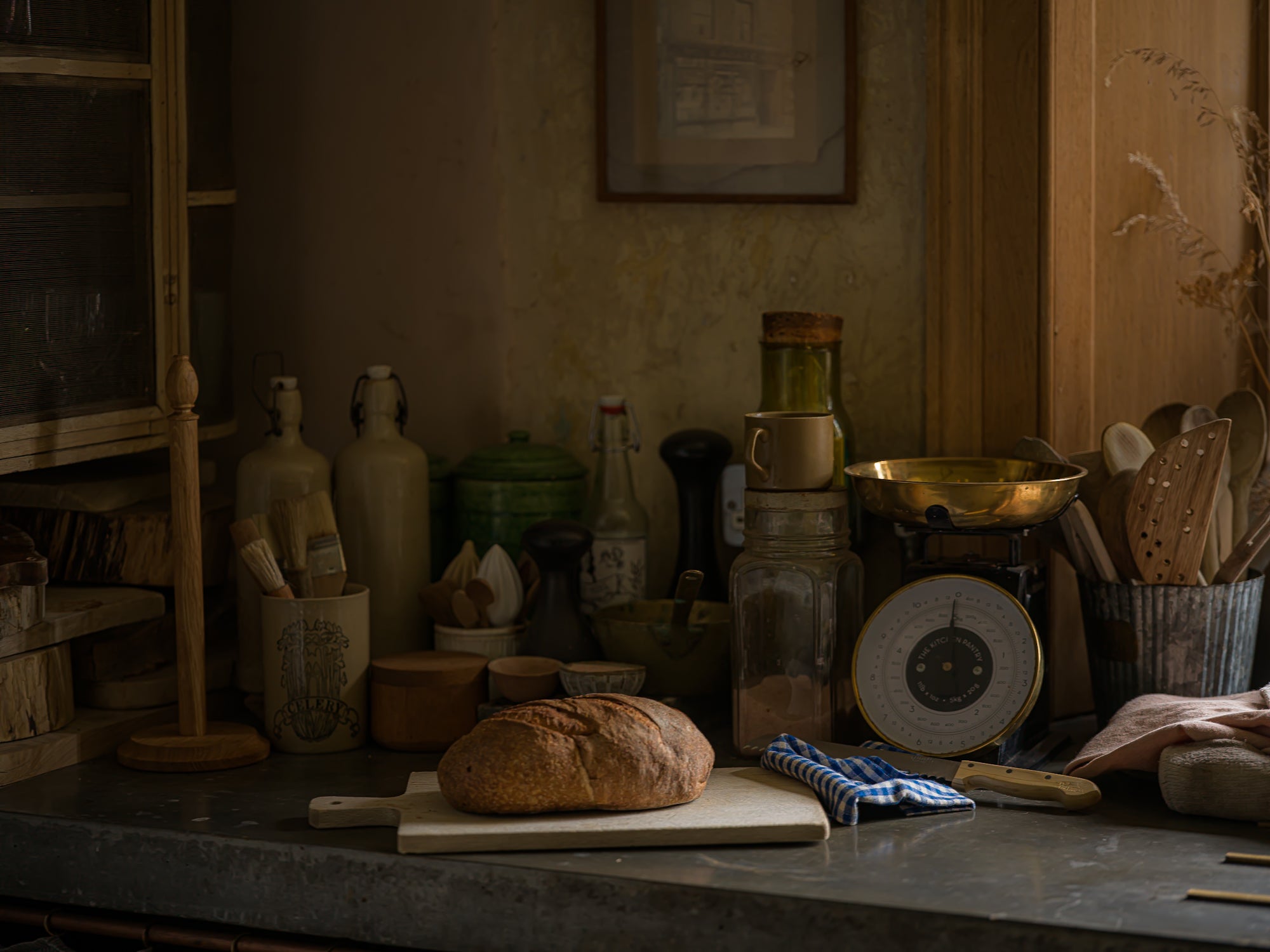 A rustic loaf of bread sits on a wooden cutting board on a dark countertop in a dimly lit, vintage-style kitchen or bakery setting. Surrounding the bread are glass bottles, jars, a brass scale, wooden utensils, and cloth towels.