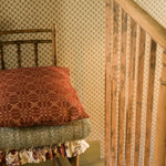 A close-up of a rustic wooden staircase railing next to a bench with a rust-red patterned throw pillow and a folded green textured blanket, set against beige patterned wallpaper.