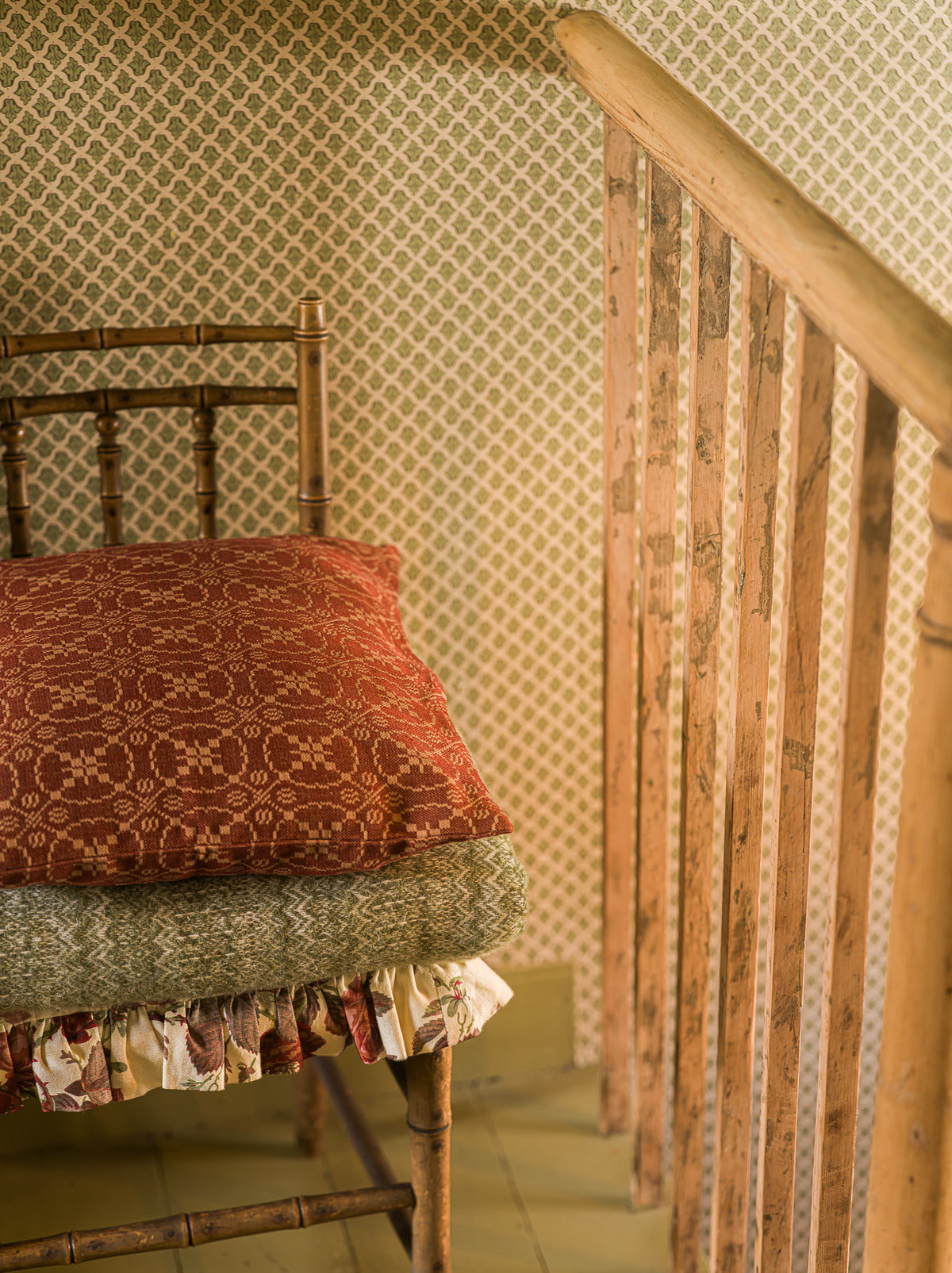 A close-up of a rustic wooden staircase railing next to a bench with a rust-red patterned throw pillow and a folded green textured blanket, set against beige patterned wallpaper.