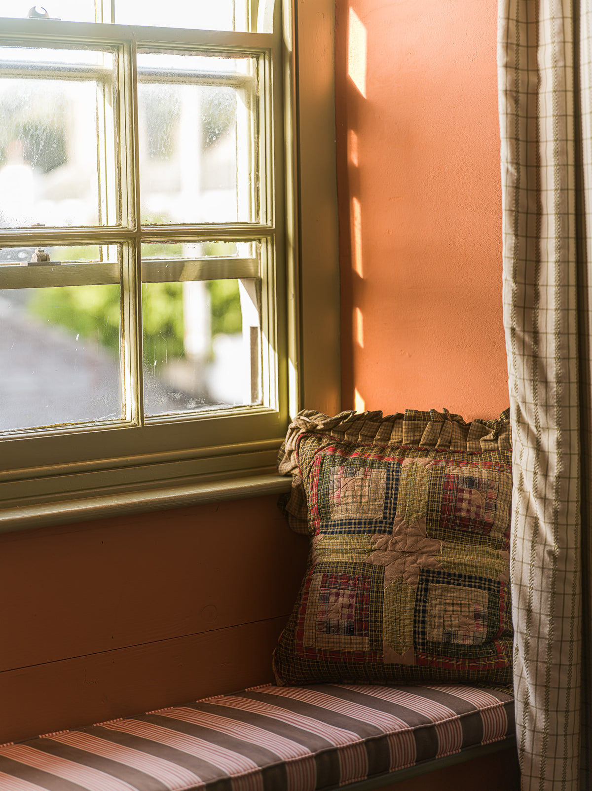 A cozy window seat cushion with a striped pink, brown, and beige cushion cover and a matching patchwork quilted throw pillow, set against a warm terracotta wall next to a window with a plaid curtain.