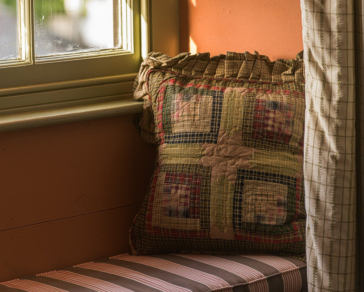 A cozy window seat cushion with a striped pink, brown, and beige cushion cover and a matching patchwork quilted throw pillow, set against a warm terracotta wall next to a window with a plaid curtain.