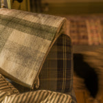 A folded plaid throw blanket in beige, gray, and muted green tones draped over the arm of a plaid upholstered chair in a warm indoor setting.
