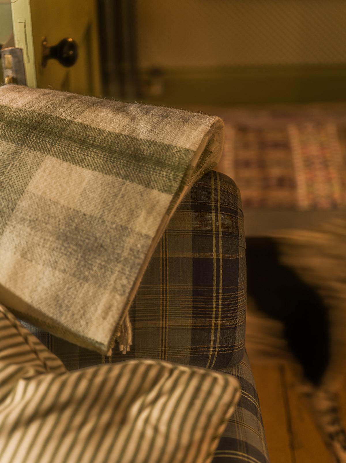 A folded plaid throw blanket in beige, gray, and muted green tones draped over the arm of a plaid upholstered chair in a warm indoor setting.