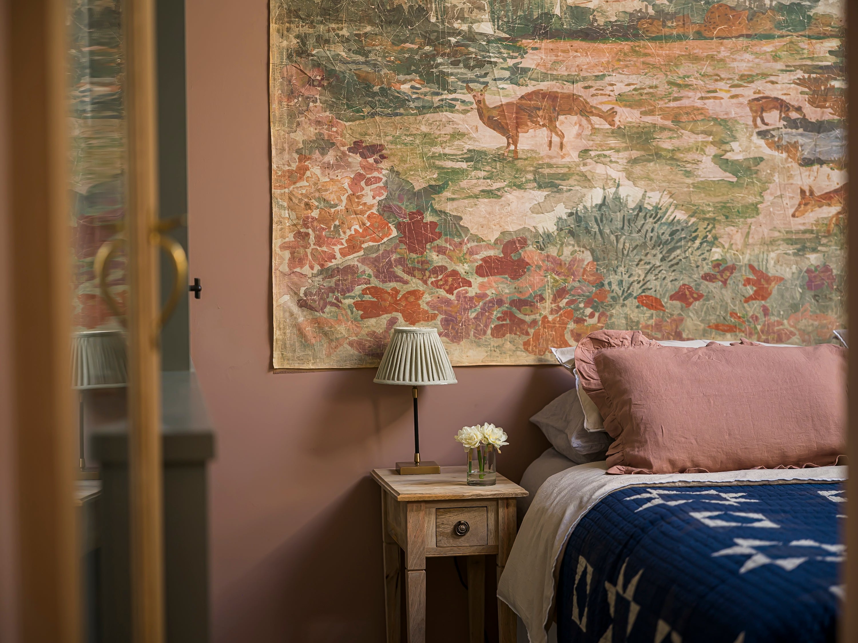 Bedroom with a large tapestry on the wall, a nightstand with a lamp and flowers, and a bed with pink pillows and a blue blanket.