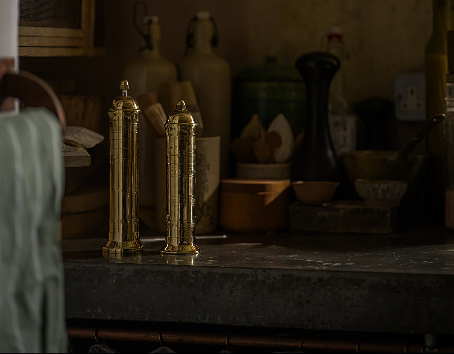 Two brass pepper mills on a wooden surface with a blurred background of pottery and decor.