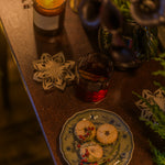 A cozy tabletop scene with a lit candle, a small glass of dark red beverage, and a plate of cookies decorated with dried flowers and berries, surrounded by holiday ornaments and greenery.