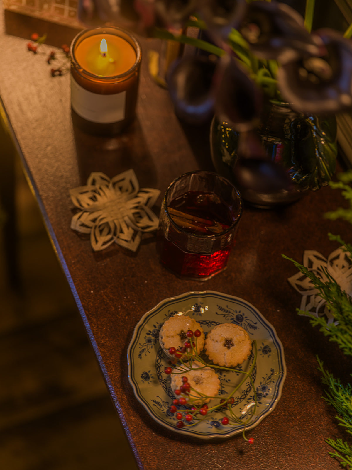 A cozy tabletop scene with a lit candle, a small glass of dark red beverage, and a plate of cookies decorated with dried flowers and berries, surrounded by holiday ornaments and greenery.