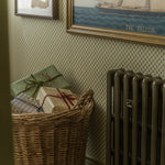A wicker basket filled with wrapped presents sits against a patterned wallpaper wall next to a dark radiator, with framed wall art visible above.