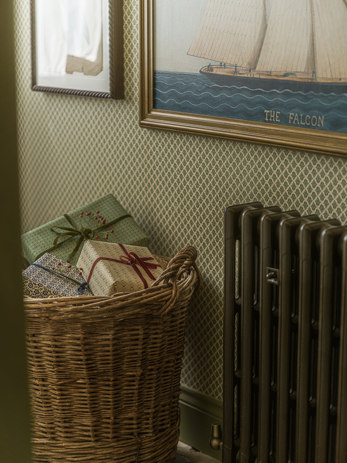 A wicker basket filled with wrapped presents sits against a patterned wallpaper wall next to a dark radiator, with framed wall art visible above.