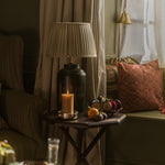 A styled interior scene with a dark wood side table holding a lit candle, a pleated table lamp, and decorative objects, set beside a window with striped curtains and a patterned cushion.