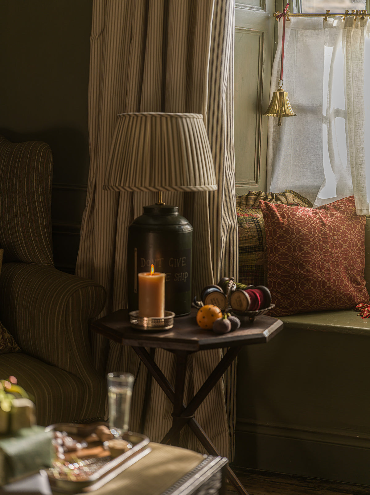 A styled interior scene with a dark wood side table holding a lit candle, a pleated table lamp, and decorative objects, set beside a window with striped curtains and a patterned cushion.
