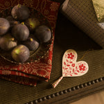A heart-shaped wooden ornament with red floral patterns hangs on a dark wooden table next to a bowl of figs and a patterned red cloth.
