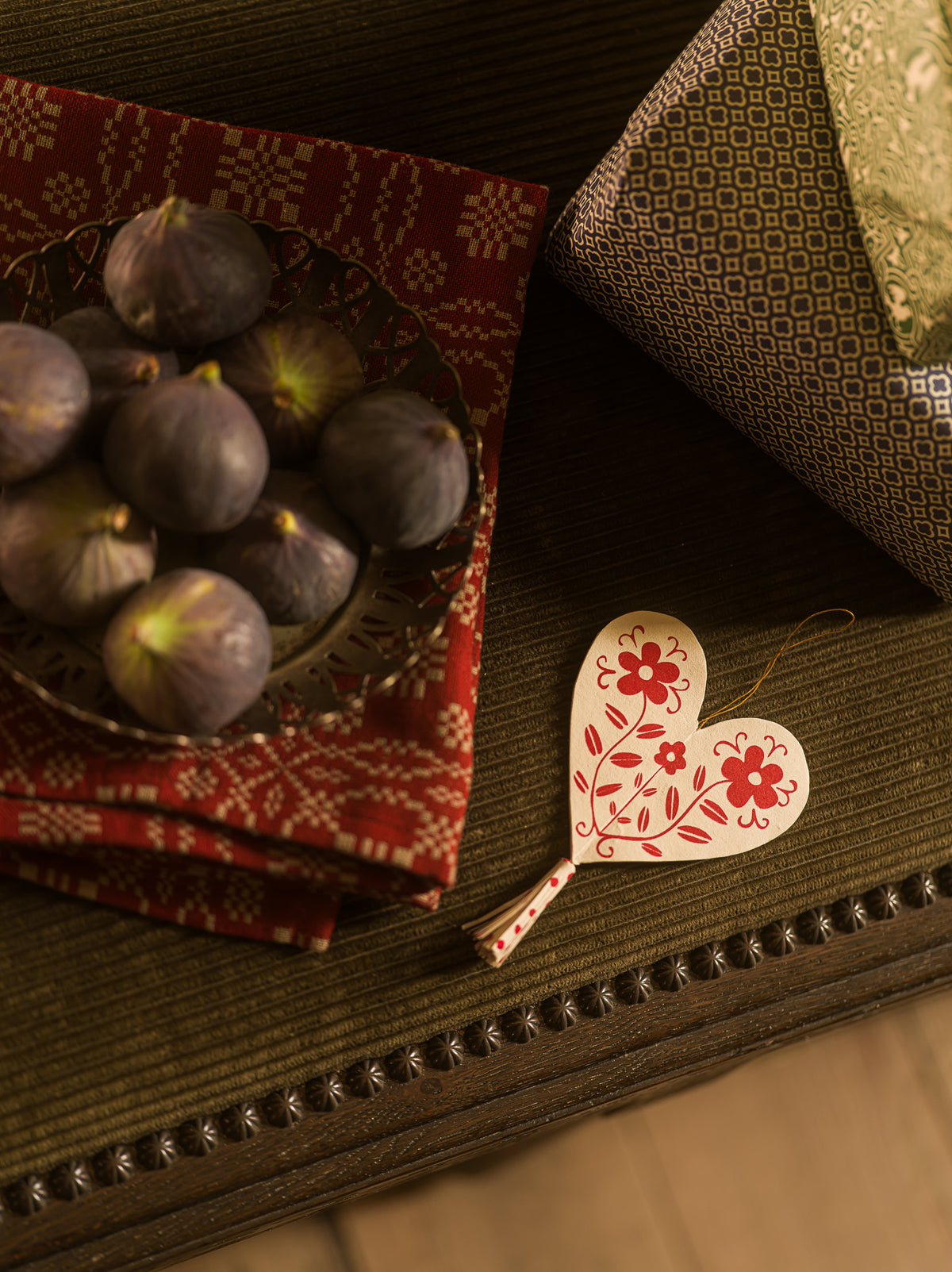 A heart-shaped wooden ornament with red floral patterns hangs on a dark wooden table next to a bowl of figs and a patterned red cloth.