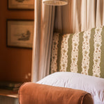 A cylindrical brown velvet lumbar pillow resting on a bed with a green and white patterned headboard, white pillows, and a warm-toned room background.