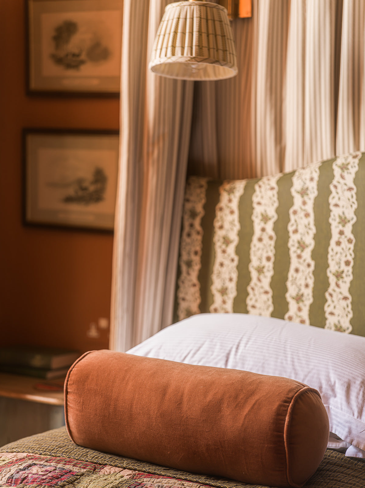 A cylindrical brown velvet lumbar pillow resting on a bed with a green and white patterned headboard, white pillows, and a warm-toned room background.