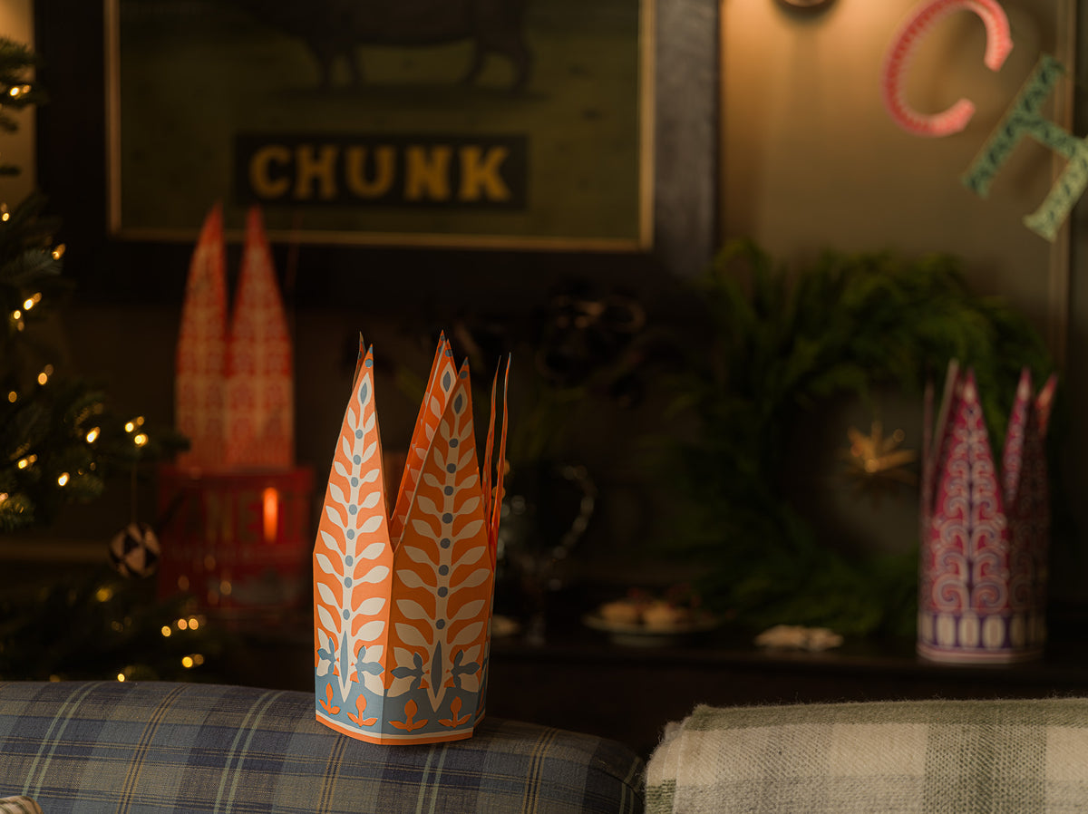 A decorative paper crown with orange and light blue leaf patterns sits on a plaid cushion in a cozy, dimly lit holiday setting with Christmas lights and a wreath in the background.