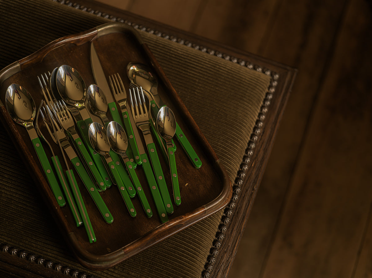 A set of cutlery with bright green handles and silver-toned metal heads, arranged on a dark tray over a brown upholstered surface.