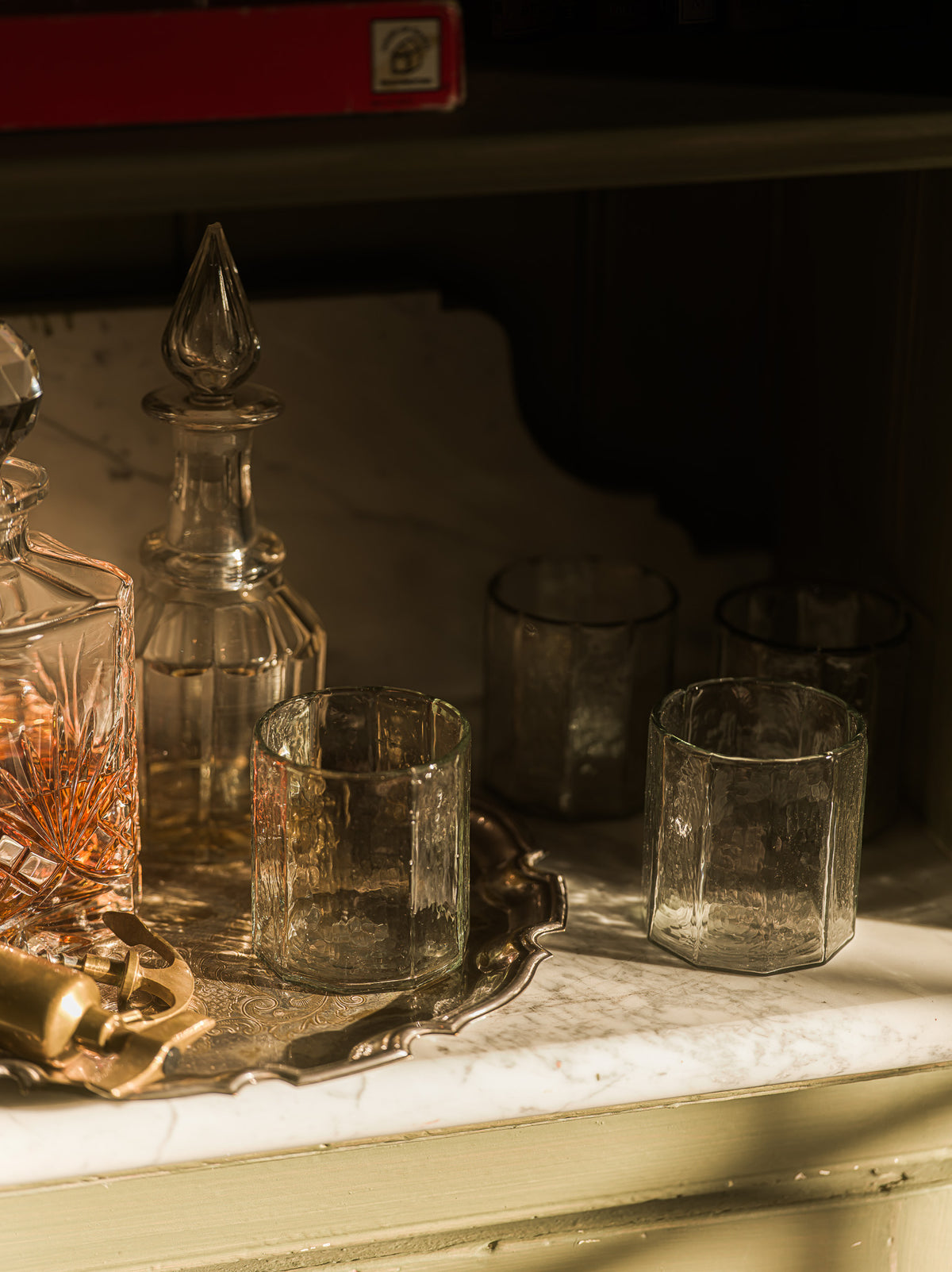A set of clear textured glass drinkingware, including tumblers and a decanter, arranged on a silver tray on a light marble surface with warm sunlight and shadows.