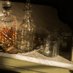 A set of clear textured glass drinking glasses and two clear glass decanters, one filled with amber liquid, arranged on a silver tray on a white marble surface.