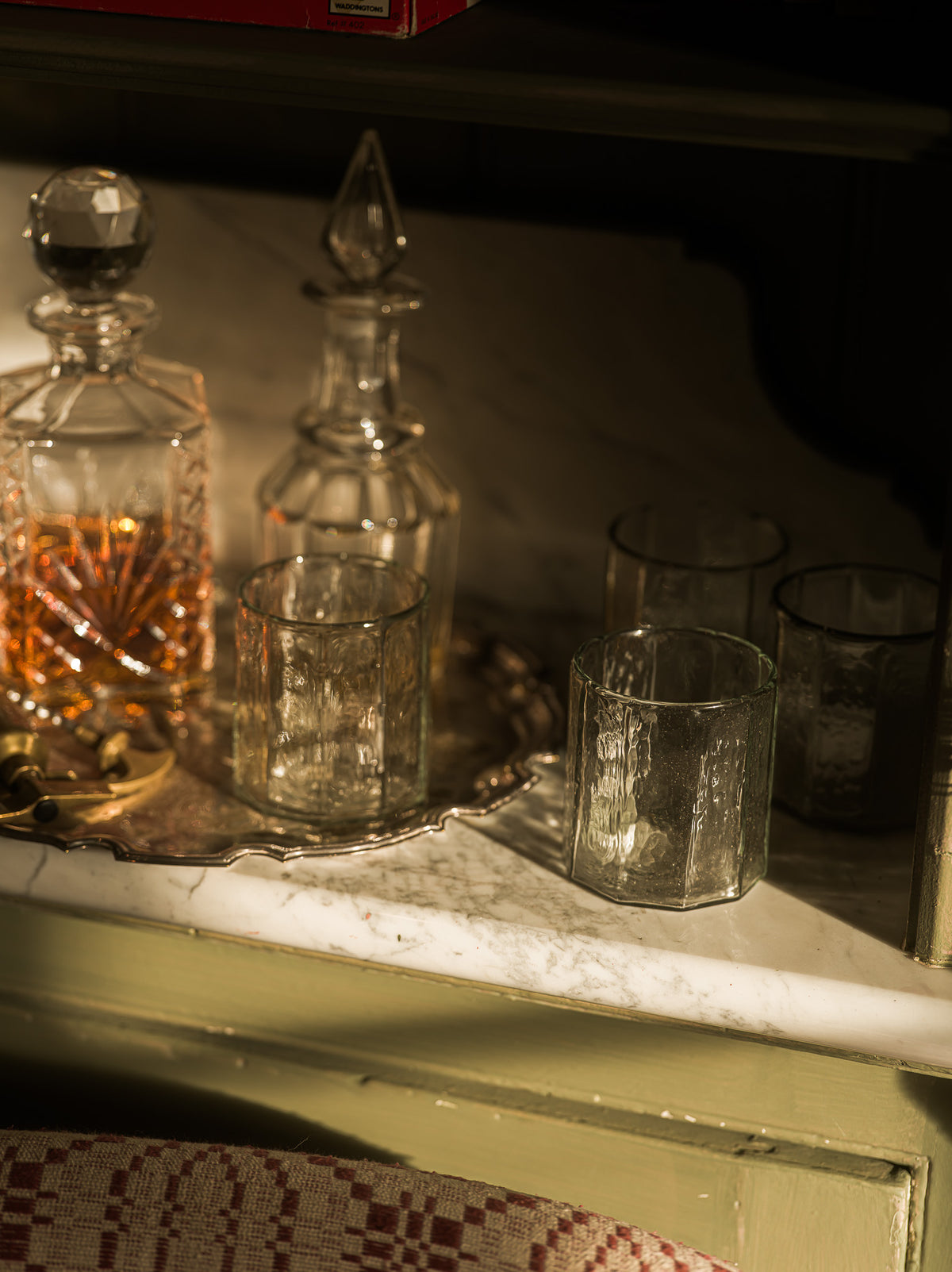 A set of clear textured glass drinking glasses and two clear glass decanters, one filled with amber liquid, arranged on a silver tray on a white marble surface.
