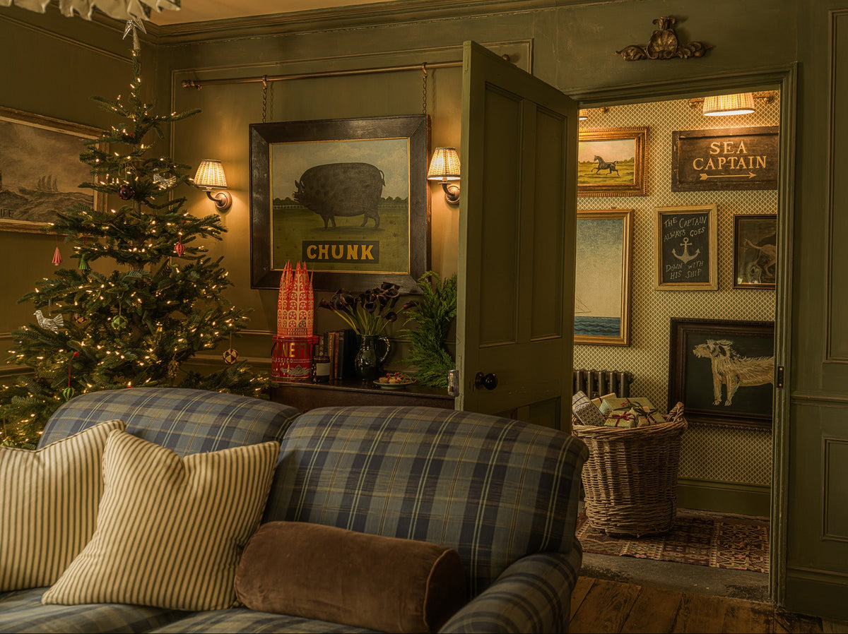 A cozy, rustic living room with dark green paneled walls, a blue and gray plaid sofa in the foreground, and an open doorway revealing a gallery wall with framed vintage-style prints including 'Chunk' and 'Sea Captain'.