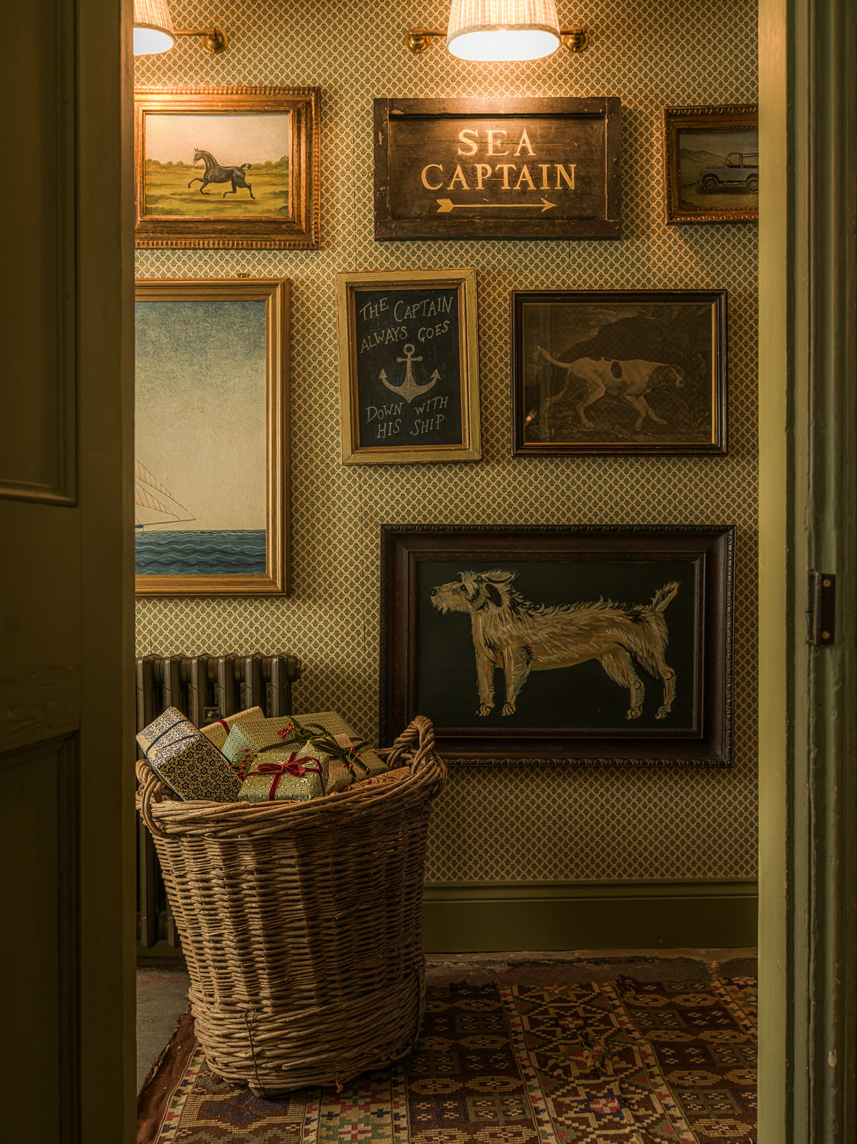 A styled interior hallway with vintage framed artwork on a patterned wallpaper wall, including a 'SEA CAPTAIN' sign, an anchor plaque, and a dog print. A large woven basket filled with books sits on a patterned rug.