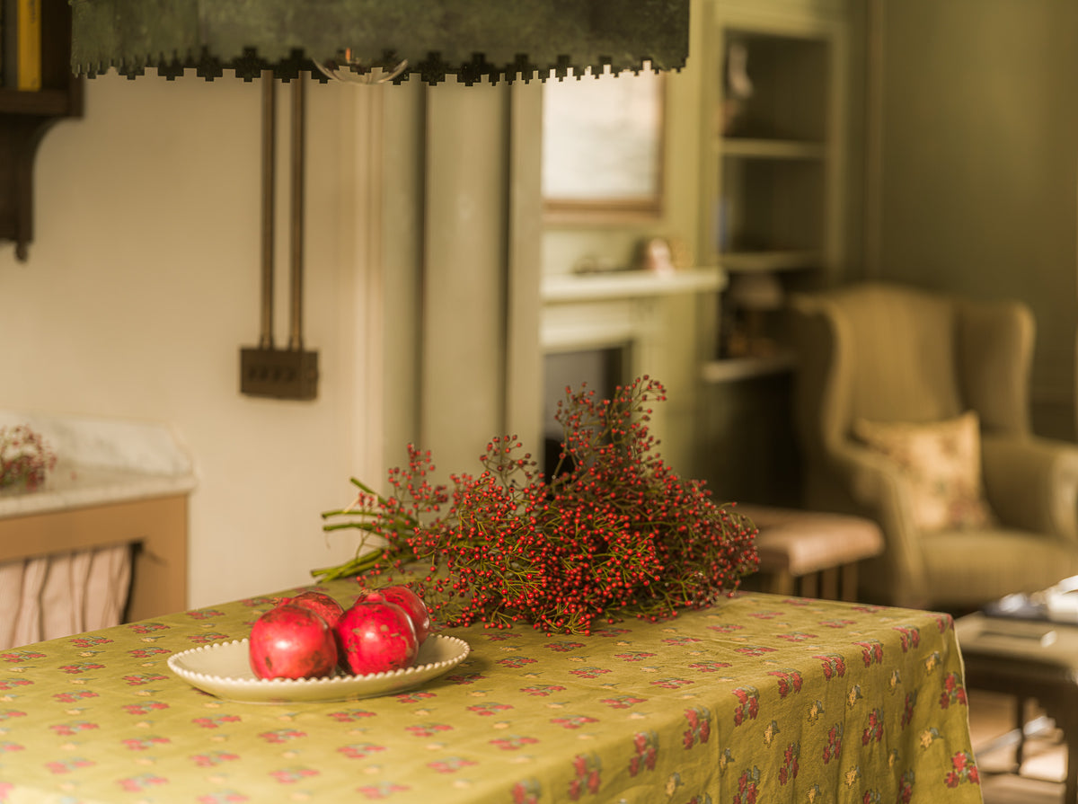 A close-up of a table covered with a yellow-green floral tablecloth, holding a white plate with three red apples and a bunch of red berries, set in a softly blurred living room interior.