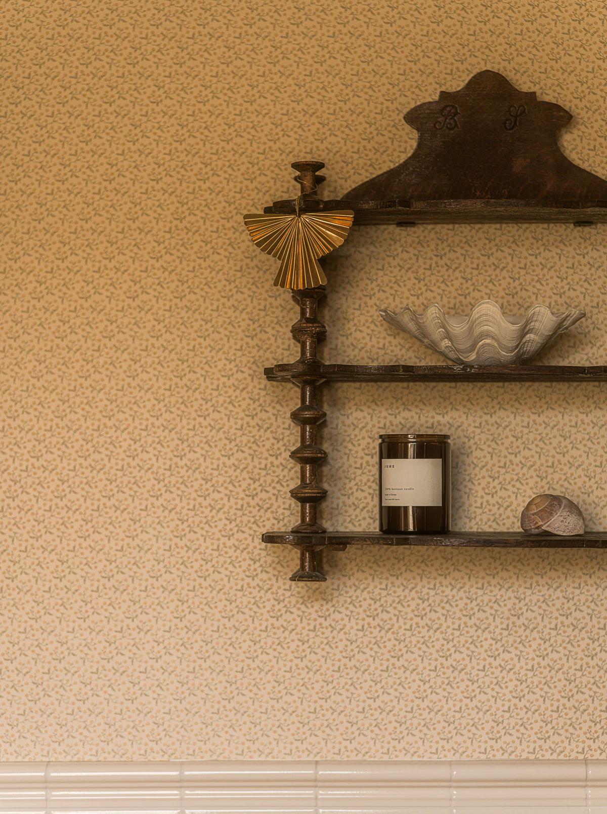 A dark brown wooden wall-mounted shelf with three tiers, styled with home decor items including a gold fan, a white seashell bowl, a candle, and another shell, set against a textured beige wallpaper background.