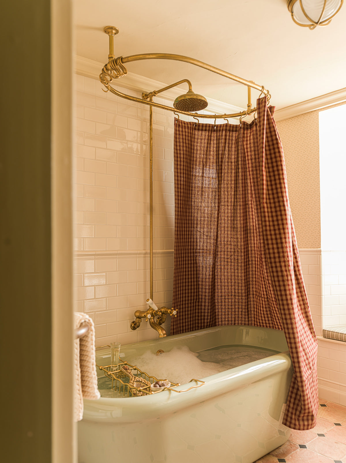 A vintage-style bathtub in a warm-toned bathroom with a rust-brown gingham shower curtain, brass shower rod and faucet, white subway tile, and a window on the right.