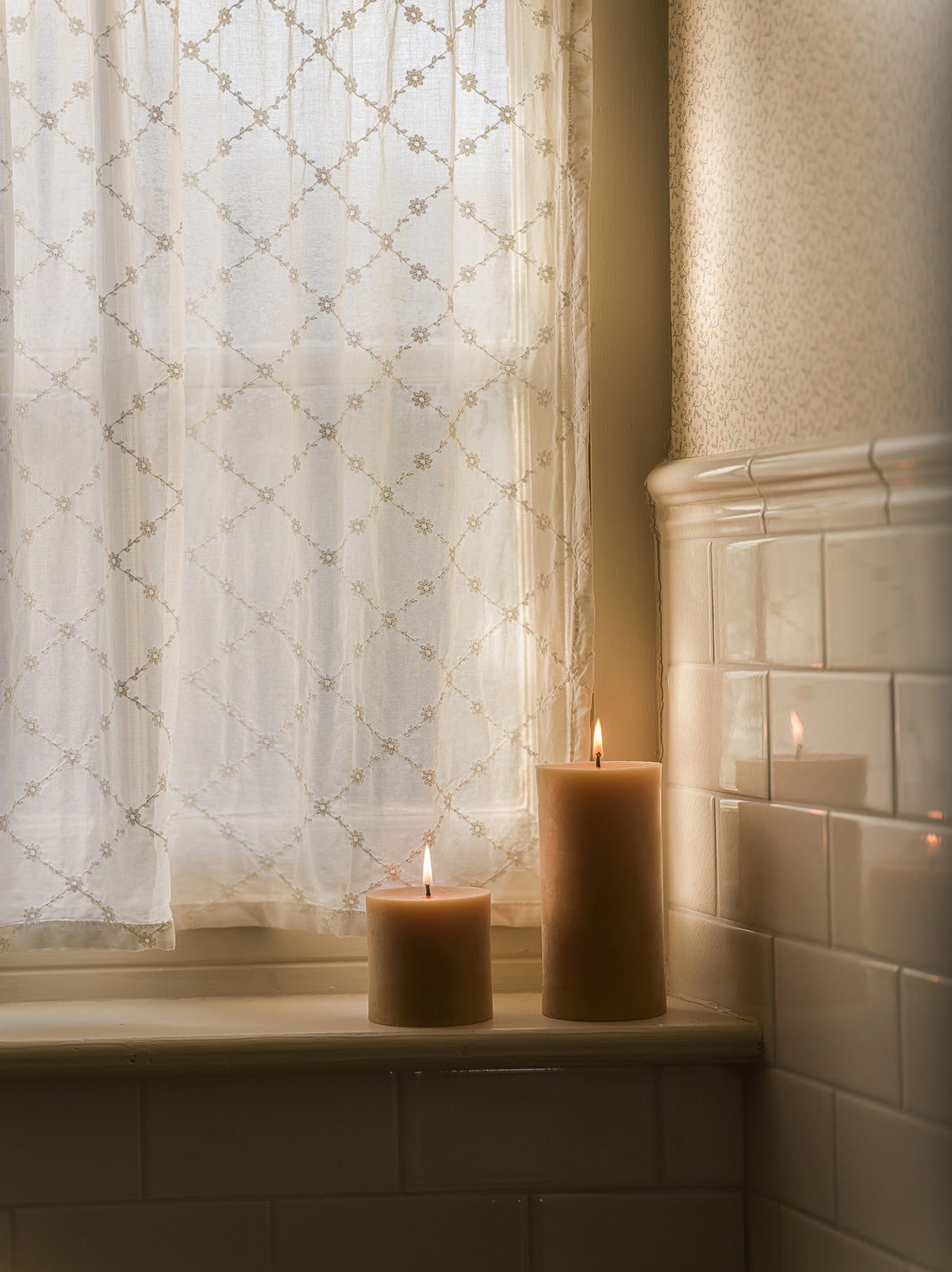Two lit beige pillar candles sit on a windowsill in a softly lit room with sheer patterned curtains, white subway tile, and warm ambient light.