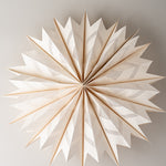 A radial, starburst-shaped decorative wall hanging made of layered white paper or fabric panels with a warm beige/tan edge, photographed against a plain light gray wall.