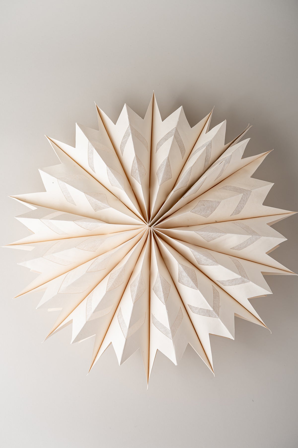 A radial, starburst-shaped decorative wall hanging made of layered white paper or fabric panels with a warm beige/tan edge, photographed against a plain light gray wall.